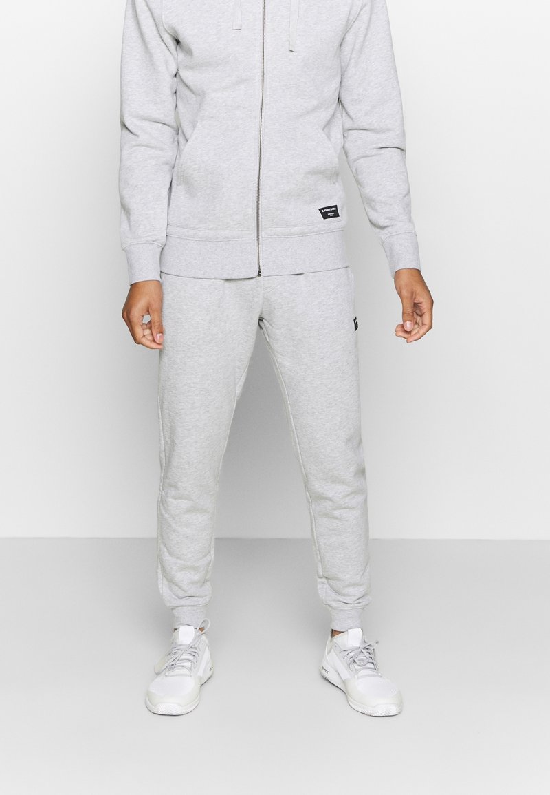Grauer Baumwollzipper mit Fronttaschen und gerippten Bündchen, kombiniert mit passenden grauen Jogginghosen. Weiße Sneaker runden das Outfit ab.