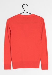 Korallfärgad stickad tröja med rund halsringning, långa ärmar och ribbad nederkant. Slät textur och klassisk passform, hängande på en svart krok.