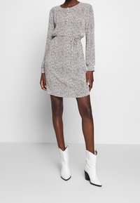 Šaty s dlouhým rukávem v černobílém puntíkovaném vzoru, s kulatým výstřihem, nařaseným pasem, doplněné bílými kotníkovými botami ve stylu kovbojských bot.