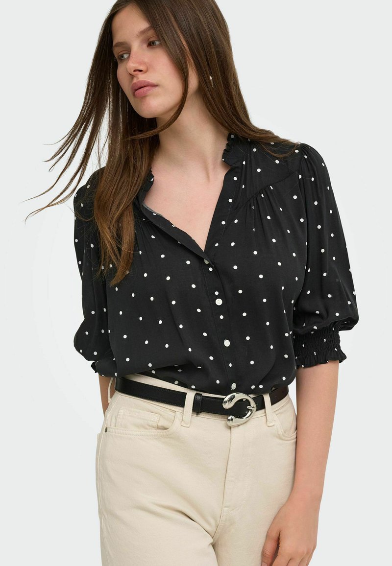 Femme portant un chemisier noir à pois blancs avec des manches trois-quarts, associé à un pantalon beige clair et une ceinture noire.