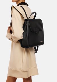 Mochila de cuero negro con cierre de solapa, superficie texturizada, herrajes decorativos y correas ajustables, llevada en la espalda de una persona.