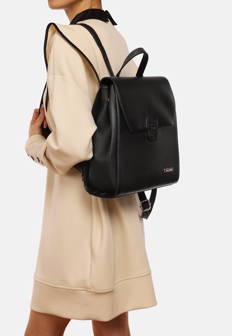 Mochila de cuero negro con cierre de solapa, superficie texturizada, herrajes decorativos y correas ajustables, llevada en la espalda de una persona.
