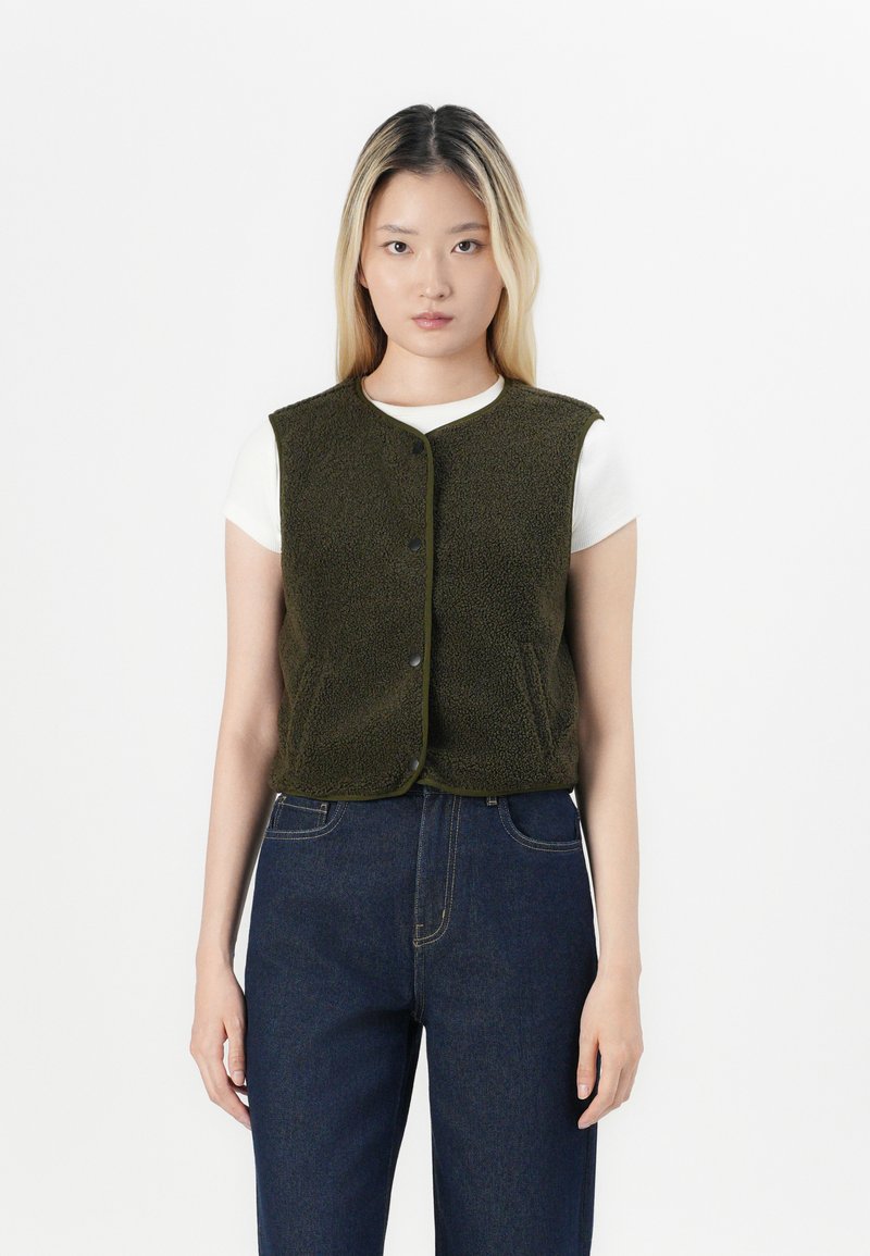 Olivenfarge fleecevest med rund hals, frontknapper og kontrastbesetning, båret over en hvit skjorte og paret med mørke jeans.
