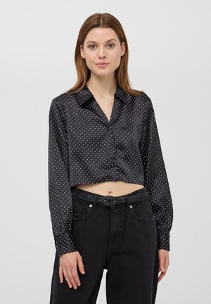 Blusa nera tagliata con pois bianchi, maniche lunghe, davanti con bottoni e colletto. Abbinata a jeans neri a vita alta e una cintura.