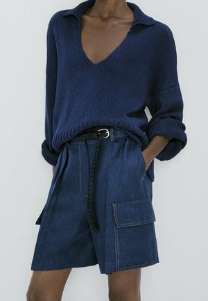 Person wearing a navy blue knit sweater with a deep V-neck and rolled sleeves, paired with high-waisted dark denim shorts with cargo pockets.