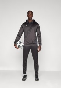 Čierna športová mikina s klokaním vreckom, zodpovedajúce čierne slim-fit nohavice a čierne tenisky. Hráč drží futbal.
