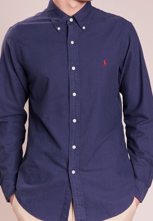 Man draagt een knoopsluitend marineblauw overhemd met lange mouwen en een rood logo op de linkerborst en beige broek, handen in de zakken.