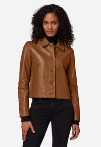 Bruine leren cropped jas met een knoopsluiting, brede kraag, lange mouwen en geribbelde manchetten. Model draagt een zwarte spijkerbroek.