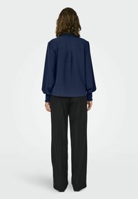 Personne aux cheveux bruns mi-longs portant un chemisier bleu marine à manches longues et un pantalon large noir, debout de dos sur un fond uni.