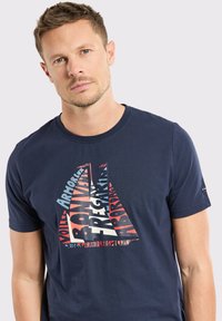 T-shirt en coton bleu marine avec un design graphique de voilier composé de texte coloré en rouge, blanc et bleu. Manches courtes et col rond.