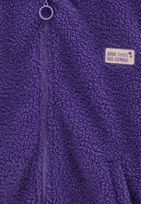Violetā flīsa jaka ar teksturētu, pūkainu materiālu. Iezīmēts ar centrālo rāvējslēdzēju un kvadrātveida etiķeti ar tekstu "LABAS LIETAS NĀK" gaiši bēšā krāsā.