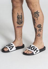Chanclas negras con una tira blanca gruesa que presenta texto en negro. La suela es negra, y la chancla deja ver los dedos y el pie al descubierto.