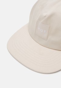 Beige kasket lavet af bomuld med en flad skygge. Har et firkantet mærke med "HUF" i hvid tekst. Tekstureret stof med synlige søm.