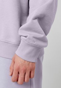 Sweatshirt lilas clair avec des poignets côtelés et un logo "BZB" ton sur ton sur la manche. Texture lisse et coupe décontractée, mettant en avant un design casual.
