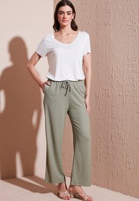 T-shirt blanc à manches courtes et col en V associé à un pantalon ample, texturé et vert clair avec une taille élastique et des jambes larges. Sandales plates visibles.