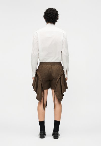 Muž s kudrnatými vlasy, který má na sobě bílou košili s dlouhým rukávem a hnědé kostičkované šortky s přehýbanými bočními panely, černé ponožky a černé boty, stojící zády.