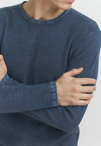 Tamno plava košulja dugih rukava s teksturiranim uzorkom, okruglim izrezom i rebrastim manžetama. Materijal izgleda mekano i prilagođeno.