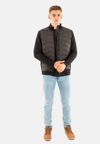 Veste matelassée noire avec un col montant, portée sur un haut à manches longues, associée à un jean bleu clair et des bottes couleur tan. Design simple avec une fermeture éclair.