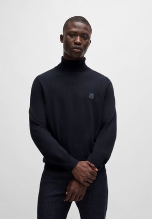 Pull à col roulé marine avec une coupe slim, des poignets côtelés et un écusson logo carré sur la poitrine. Le tissu semble lisse et doux.