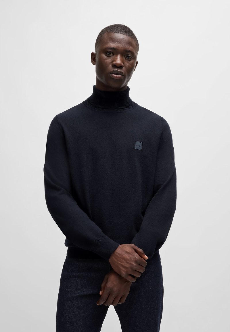 Mornarsko pulover s turtleneckom, oprijetega kroja, z ribanimi manšetami in kvadratno oznako na prsih. Tkanina se zdi gladka in mehka.