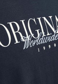 Granatowa bawełniana koszulka z wyraźnym białym i jasnoniebieskim napisem "ORIGINAL Worldwide 1990" o gładkiej teksturze.