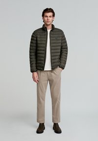 Chaqueta acolchada de color verde oliva con cremallera al frente y cuello alto; llevada sobre una camiseta blanca y combinada con pantalones beiges y botas negras.
