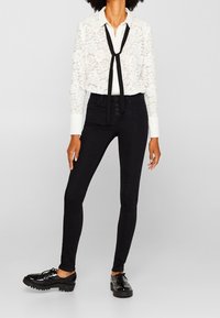 Jeans ajustados negros combinados con una blusa de encaje blanca que cuenta con una corbata negra. El conjunto incluye zapatos negros de cordones con suelas gruesas.