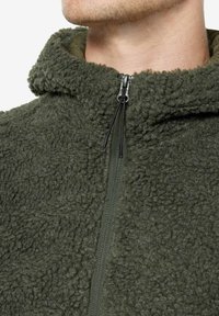 Giacca in pile verde oliva con superficie strutturata, colletto alto e chiusura con zip dotata di un fermaglio nero. Materiale morbido e caldo.