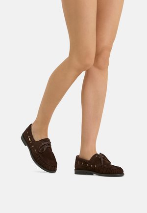 Legs wearing dark brown suede moccasin shoes with laces, standing on a light neutral background.