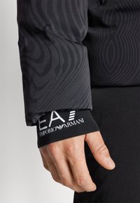 Mâneca athletică neagră cu un design texturat, prezentând linii ondulate. Poarta logo-ul și detaliile de branding "EA7 Emporio Armani" în alb.