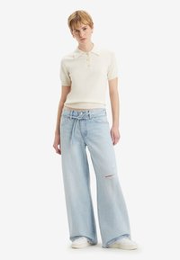 Lys beige tettsittende polo skjorte med korte ermer, parret med vide lyseblå ripped jeans og hvite sneakers.