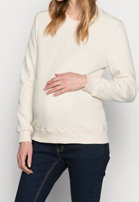 Un sweat-shirt couleur crème avec un col rond et des poignets côtelés, conçu pour une silhouette de femme enceinte, associé à un jean en denim bleu foncé.