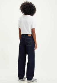 Pantalones vaqueros anchos de color azul oscuro con costuras naranjas, bolsillos traseros y una etiqueta de cuero de la marca. Combinados con una camiseta blanca de manga corta.