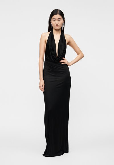 Nő ujjatlan fekete, mélyen kivágott nyakú, hosszú sziluettű ruhában, hosszú lógó fülbevalókkal, kezét a csípőjén tartva áll.