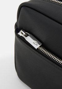 Angolo di una borsa in tessuto nero con una texture, dotata di zip e accessori in argento. Dettaglio del logo visibile sul tirante della zip. Forma compatta con linee pulite.
