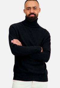 Pull en col montant noir avec des poignets et un ourlet côtelés, présentant une texture douce et une coupe décontractée, porté par une personne les bras croisés.