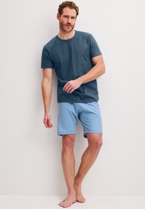 Man met krullend haar, gekleed in een blauw T-shirt en lichtblauwe korte broek met patroon, staat blootsvoets tegen een effen witte muur.