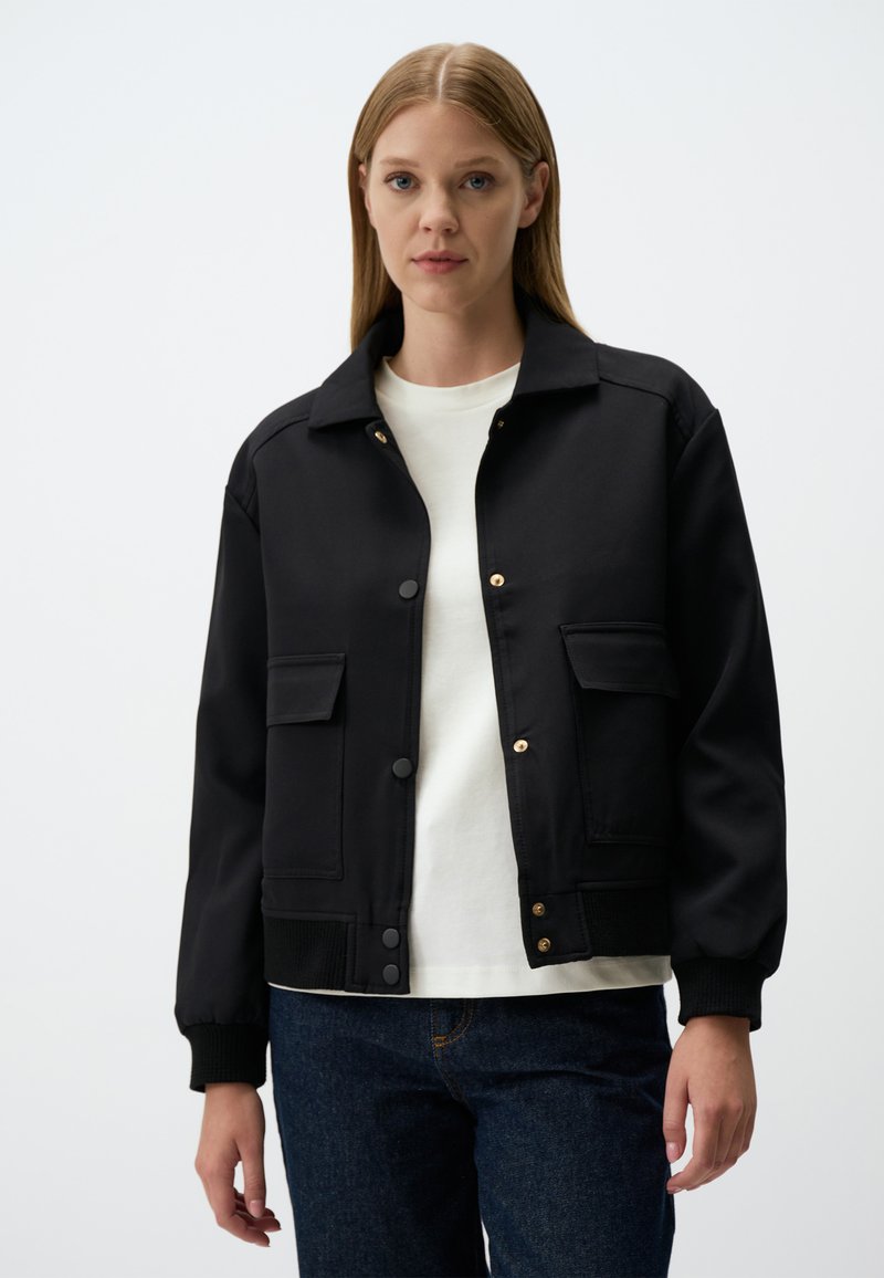 Veste bomber noire avec un col, des boutons-pression, des poches latérales, des poignets côtelés et une texture lisse. Portée par-dessus un t-shirt blanc.
