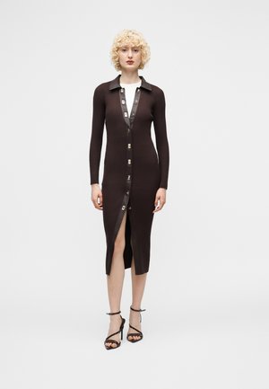 Kobieta o kręconych blond włosach, ubrana w długą, prążkowaną, ciemnobrązową sukienkę midi z guzikami z przodu oraz czarne wysokie sandały na paski.