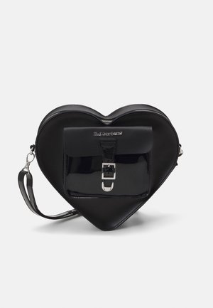 Sac à main noir en forme de cœur avec poche avant à boucle, fermeture éclair argentée et bracelet détachable sur fond blanc.