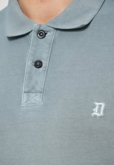 Ανοιχτό μπλε πόλο μπλουζάκι από υφασμάτινη υφή με ριμπ λαιμόκοψη, δύο μαύρα κουμπιά και μικρό λευκό κεντημένο λογότυπο 'D'.