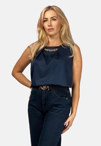 Granatowa bluzka bez rękawów z plisą z przodu i złotym łańcuszkiem przy dekolcie, zestawiona z ciemnymi dżinsami o wysokim stanie.