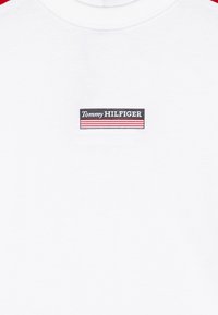 T-shirt bianco in cotone con un'etichetta nera rettangolare con "Tommy HILFIGER" e strisce rosse, centrata sul petto. Maniche corte.