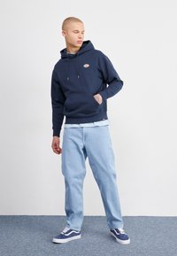 Felpa navy con tasca frontale e cappuccio con coulisse, abbinata a jeans azzurri e scarpe da ginnastica blu scuro con accenti bianchi.