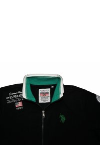 Svart zip-jacka med en grön innersnarv, vita randiga detaljer, broderad logotyp och tryckt text på vänster bröstområde.