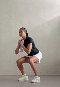 Osoba čuči držeći srebrnu bučicu, odjevena u crnu majicu i bijele šorceve, s zelenim i bijelim sportskim cipelama.