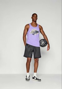 Homme grand portant un débardeur violet des Lakers, un short de basket noir et des baskets, tenant un ballon de basket Spalding noir devant un fond uni.