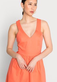 Vestido sem mangas coral com decote em V profundo, corpete justo e cintura franzida. Tecido suave e leve com saia levemente fluida.