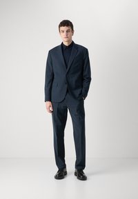 Abito blu navy con giacca monopetto, rever a scialle, pantaloni con frontale piatto e scarpe nere. Tessuto liscio con vestibilità su misura.
