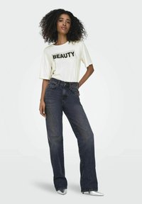 Witte t-shirt met "BEAUTY" in zwarte letters, gecombineerd met donkerblauwe hoge taille jeans en zilveren stiletto hakken. Krullend haar.